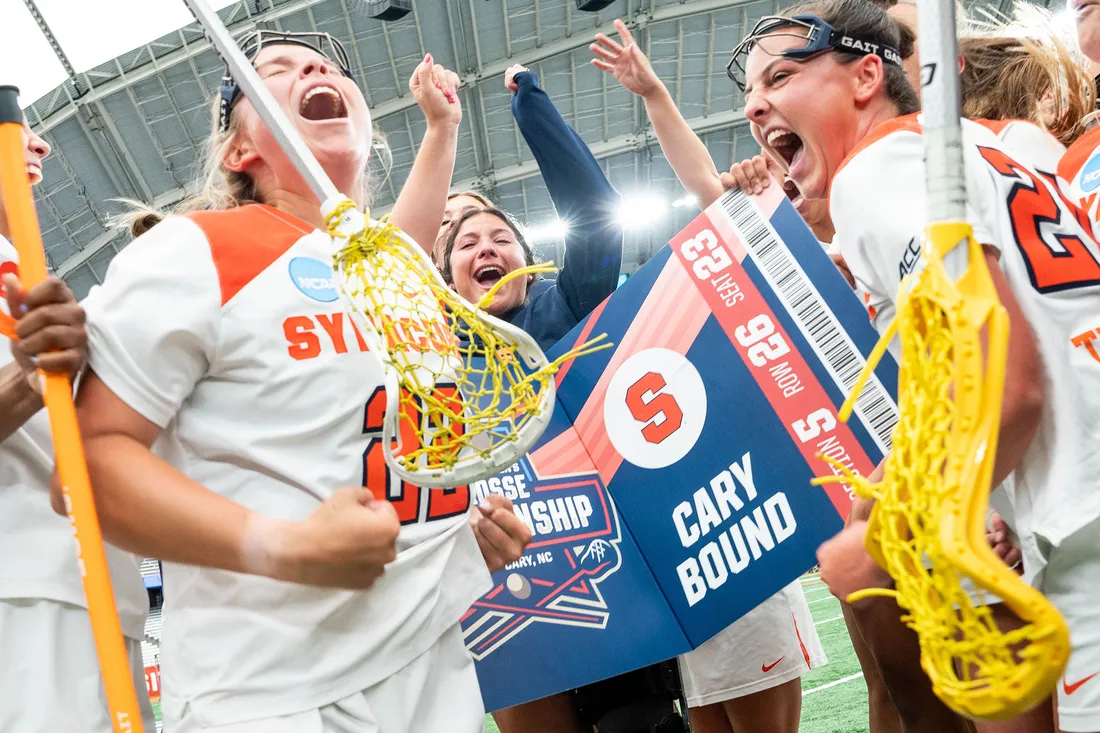 Women's lacrosse team celebrates moving on to national championship on the field holding a banner with "Cary Bound" written on it.