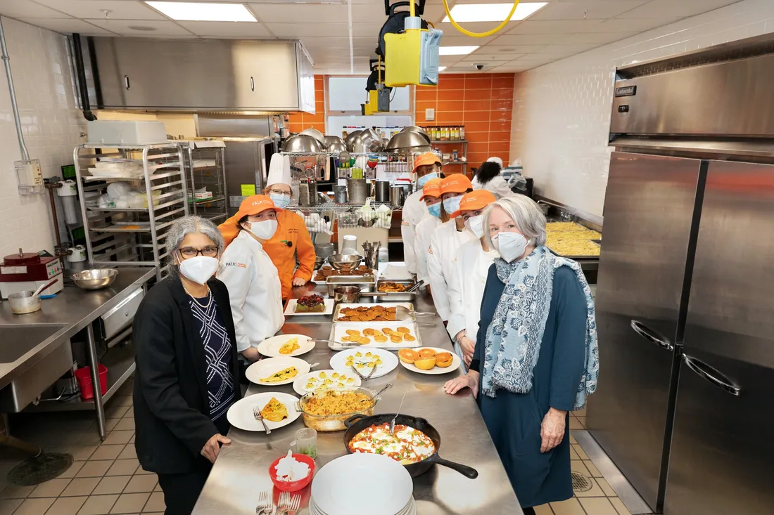 View of people standing in commercial kitchen next to table covered with dishes of food.