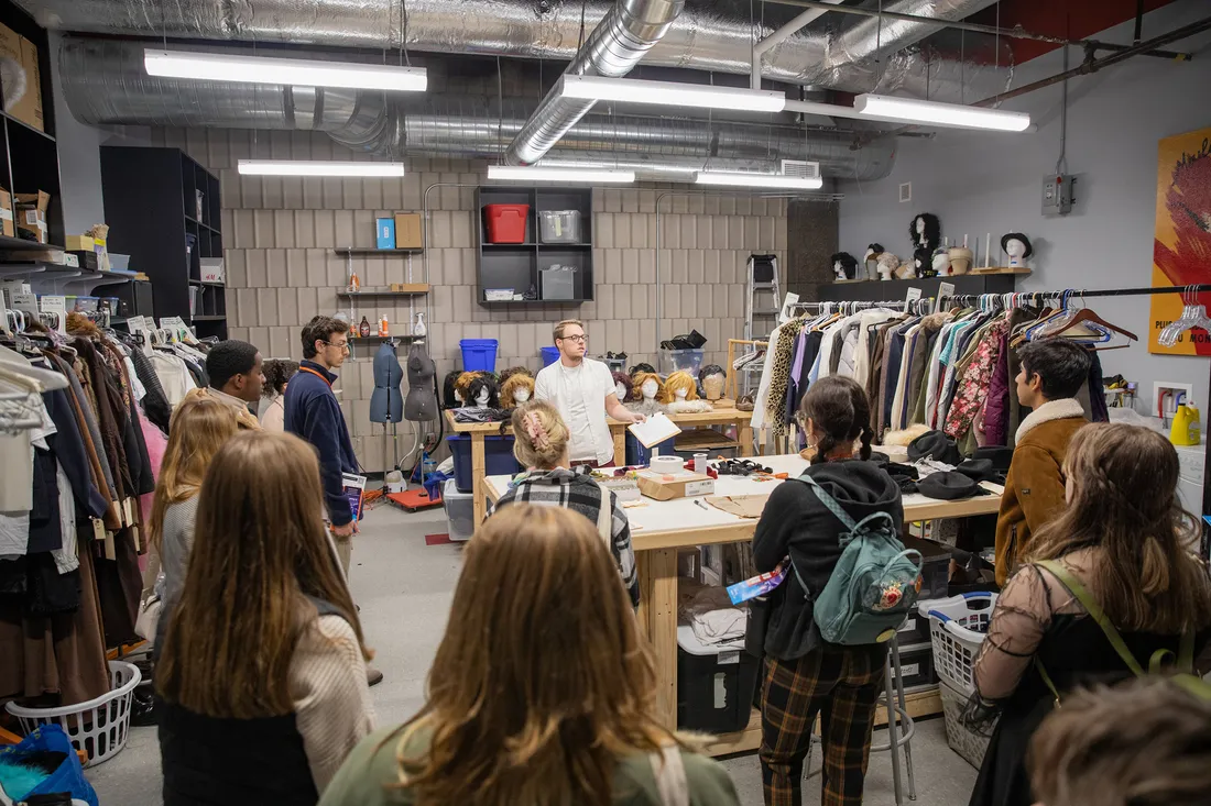 Photo of students gathering around a worktable, surrounded by clothing racks, wigs and other costuming.