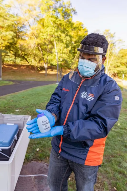 Person wearing mask, face shield and gloves shows the camera a water sample in a container.