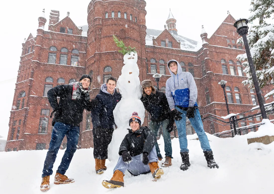 five people around a tall snow sculpture on a snowy hill with a brick building in the background