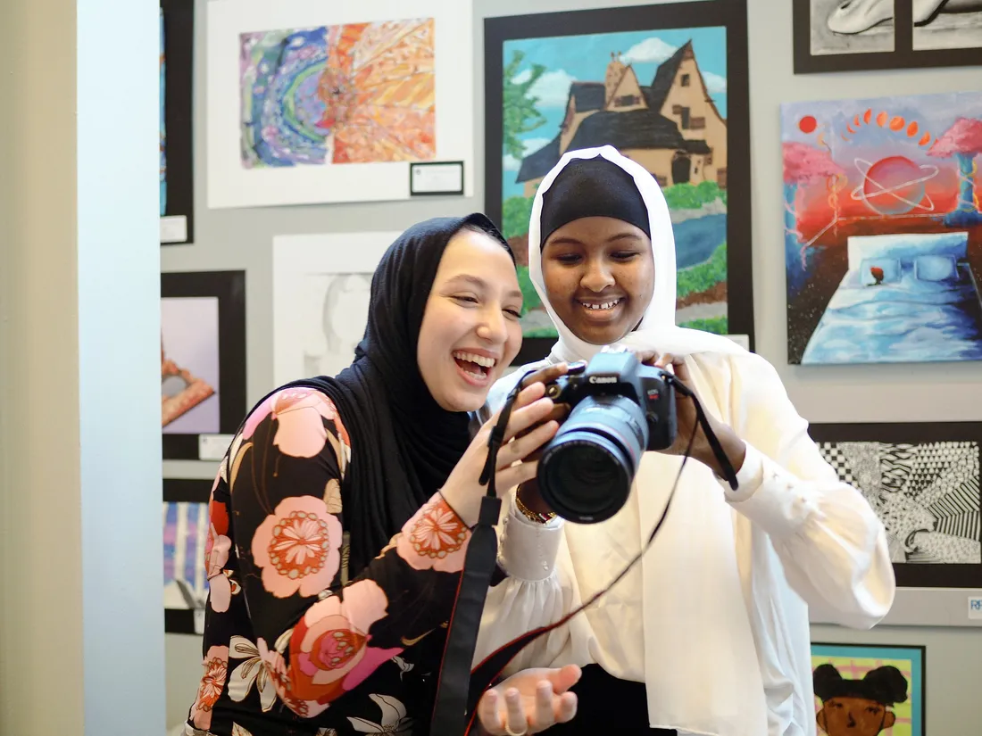 Two smiling young women stand side by side looking into a camera.