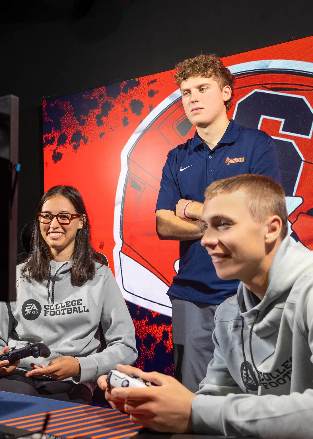 Two students playing a video game, and one student standing above them watching.