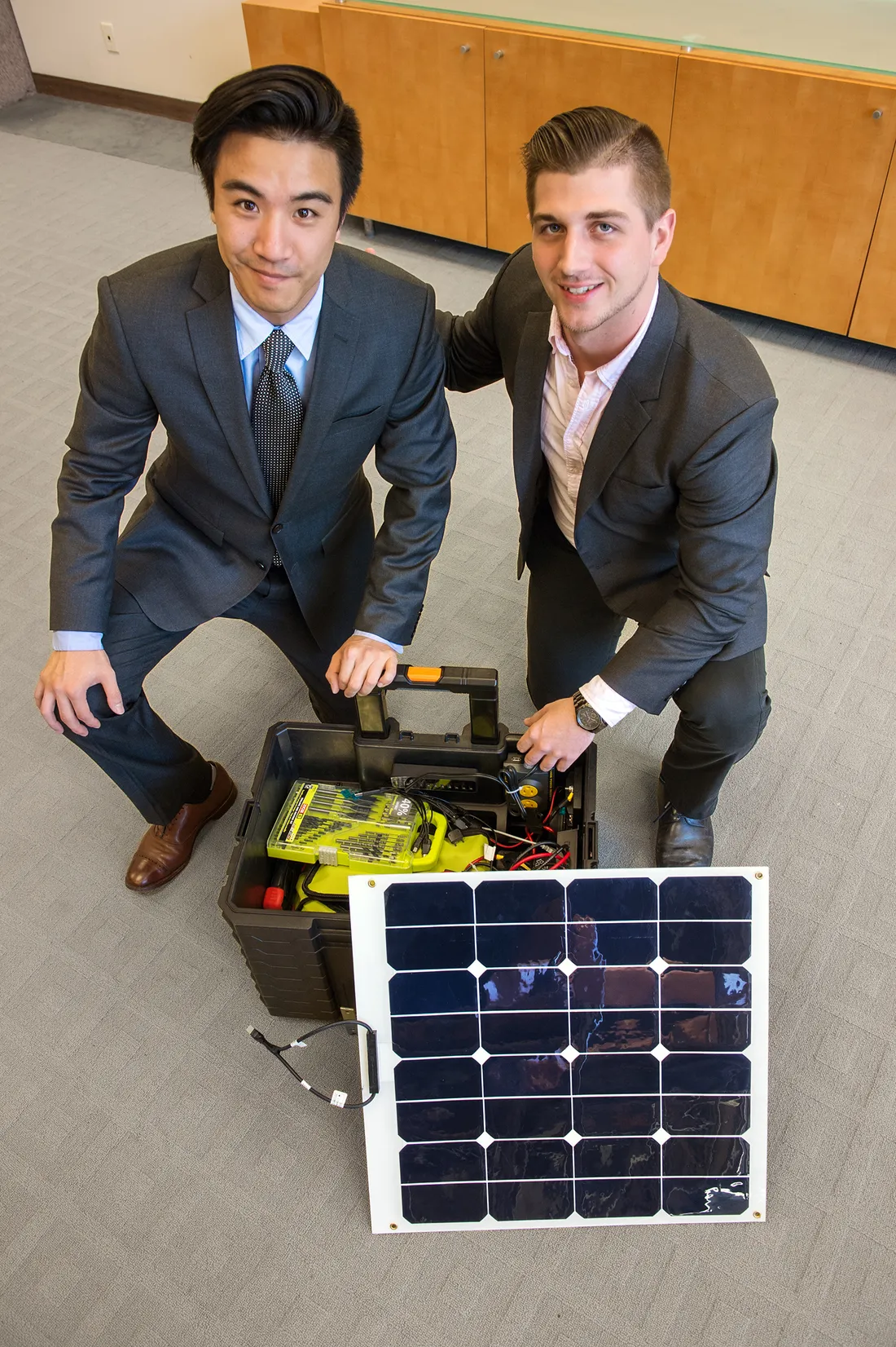 Two men sit by small solar panel.