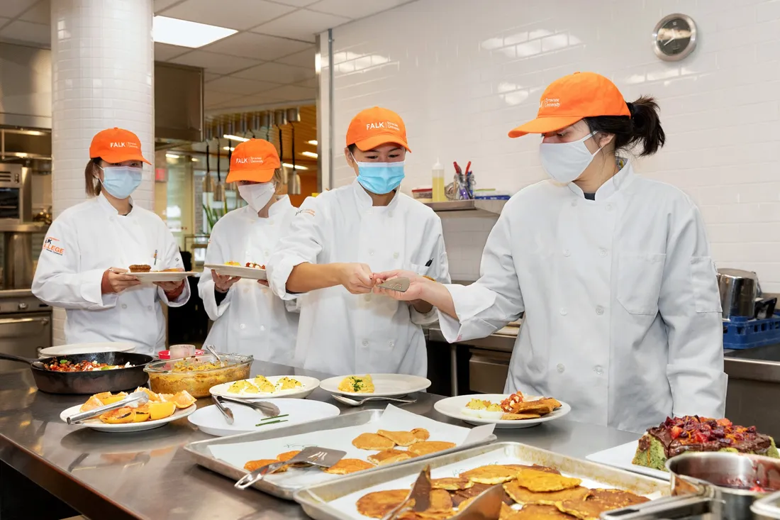 Four students in chef coats stand behind a table covered with dishes of food.
