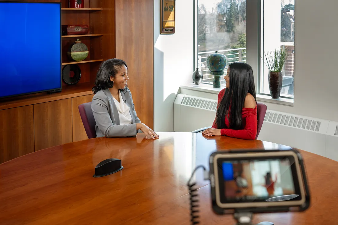 Two people sitting at table talking, while recording a video.