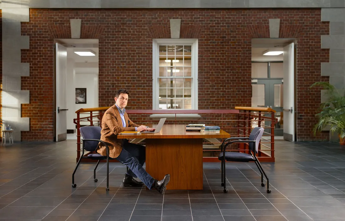Professor posing at a desk in front of a brick building.