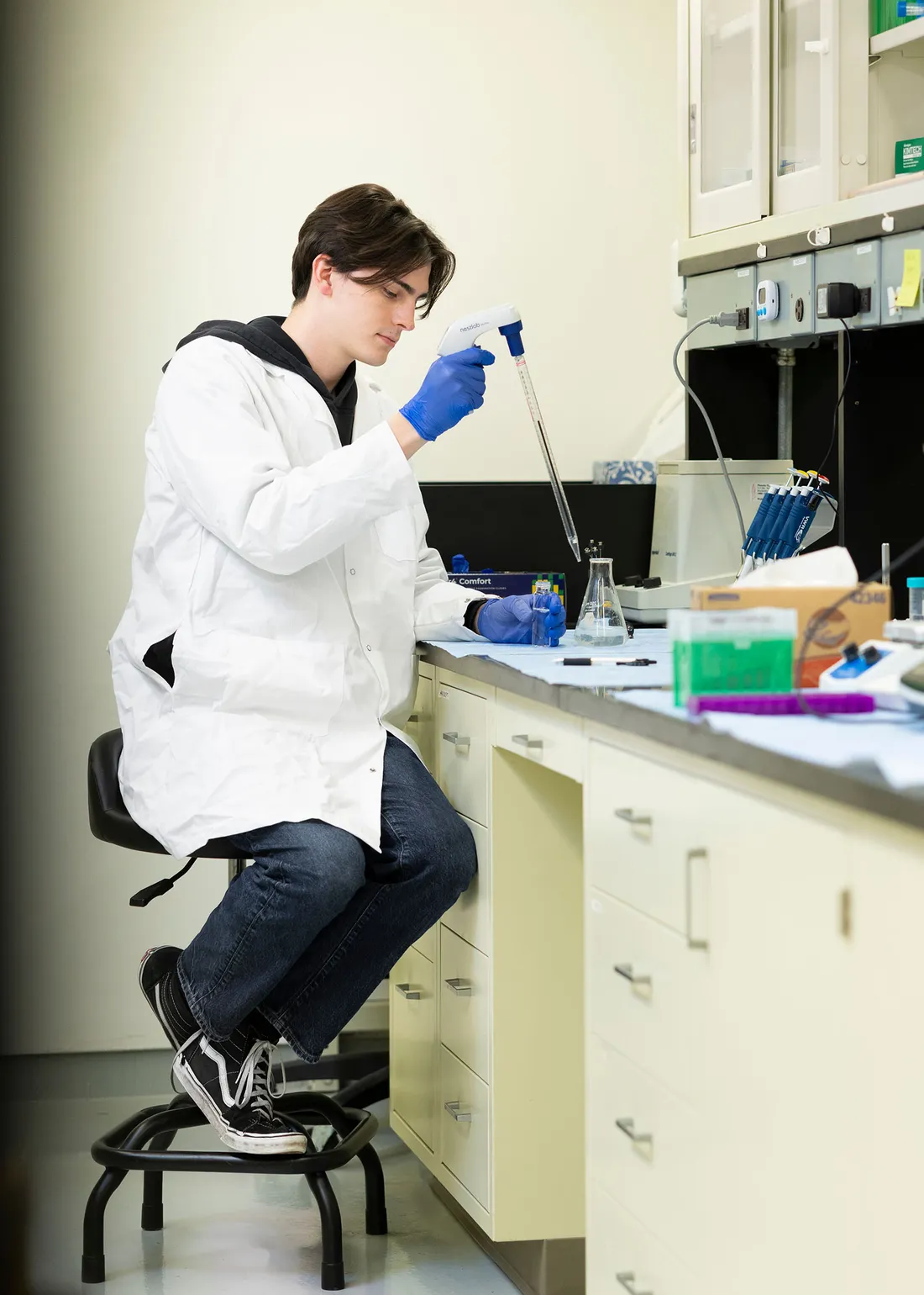 A student in a white lab coat sitting on a bench and holding a pipet.