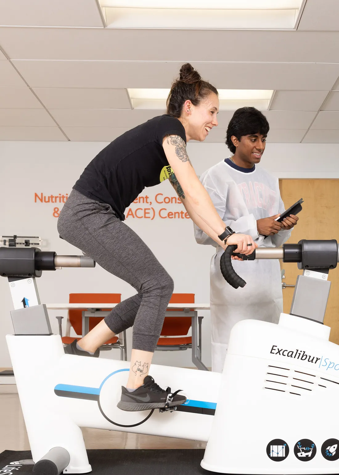 Person on a stationary bike and a person recording data next to her.
