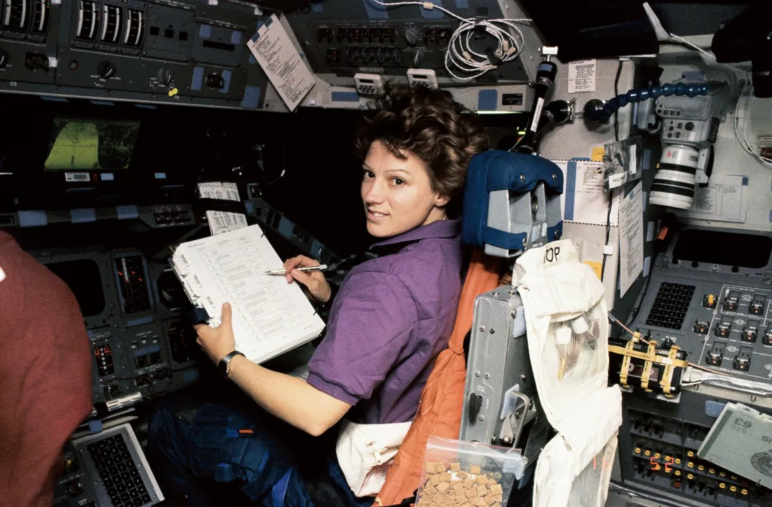 Eileen Collins in a space shuttle, with a clipboard and checklist.