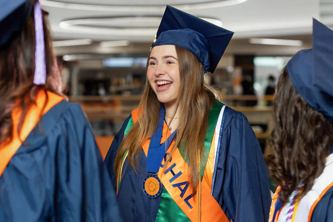 Rylee Pepper standing and smiling and talking with a friend in their commencement regalia.