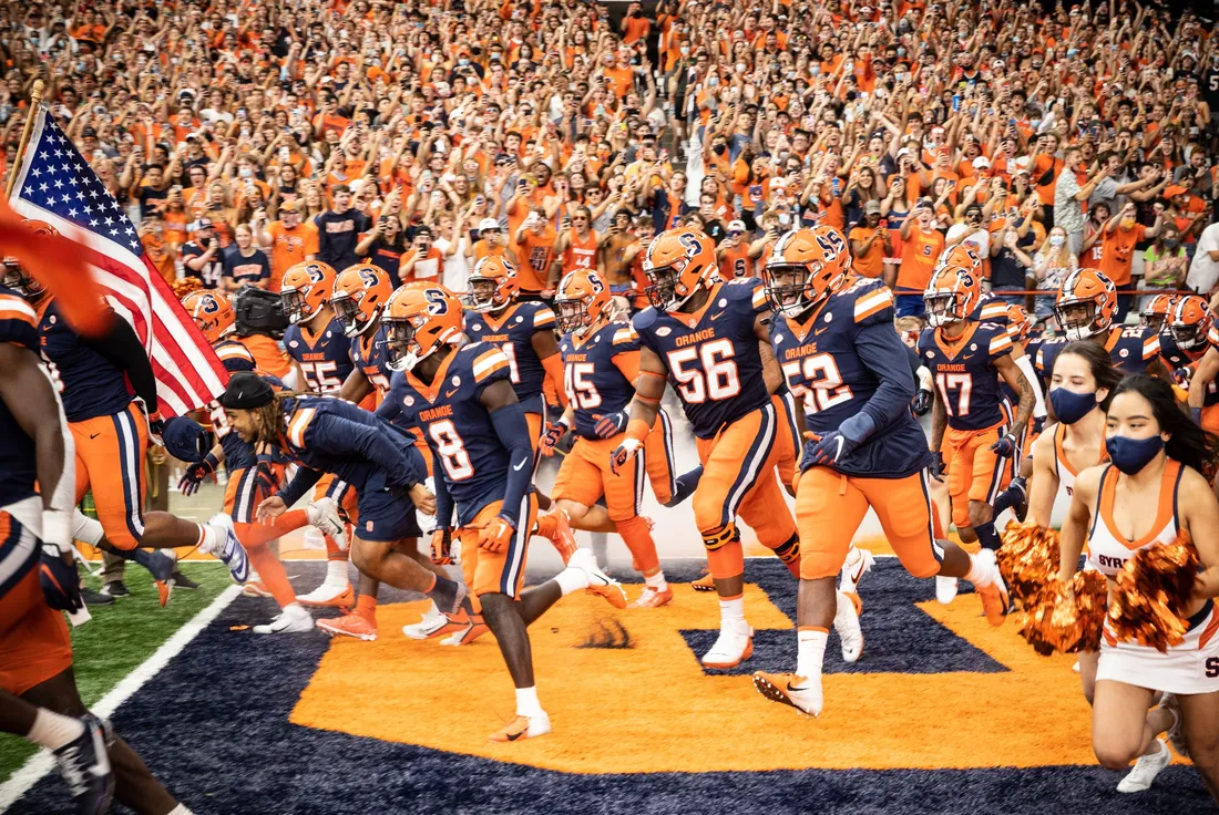 Syracuse University football players and cheerleaders running into stadium, with cheering fans behind them.