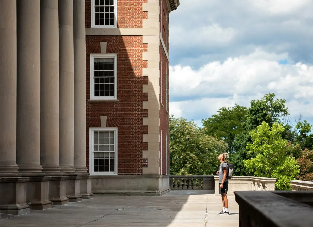 An individual stands in front of the main entrance to the Maxwell School of Citizenship and Public Affairs at Syracuse University.