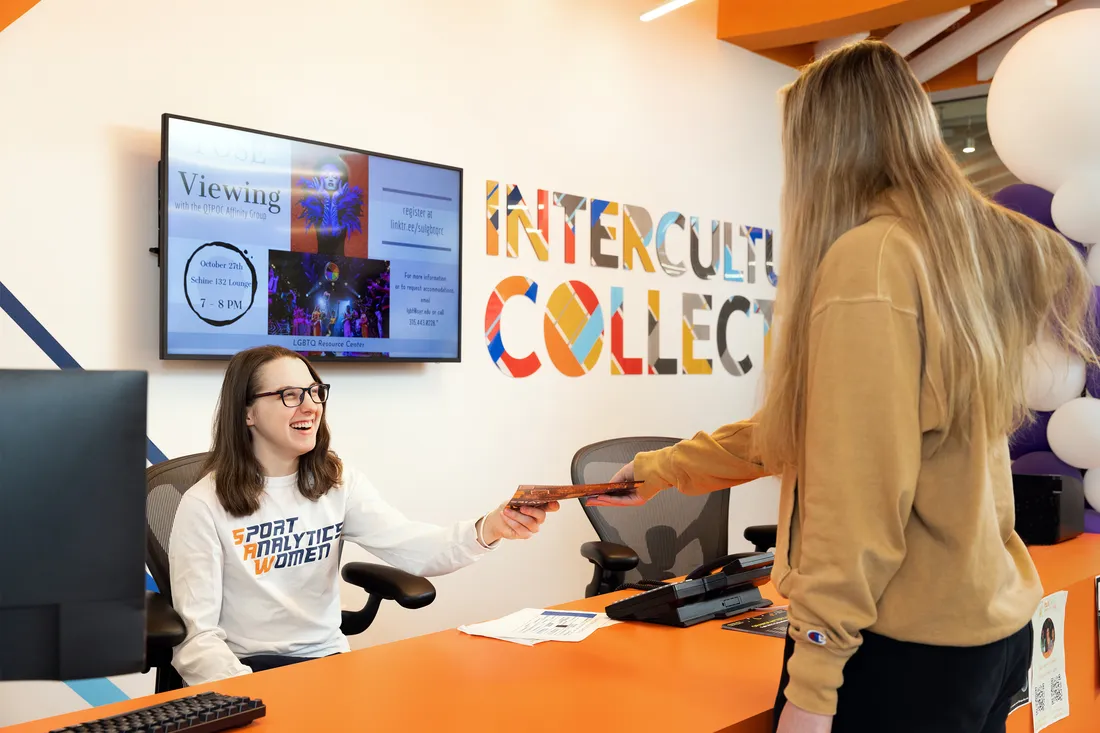 Photo of Gilmore sitting behind a desk at the Intercultural Collective, smiling and handing a student.