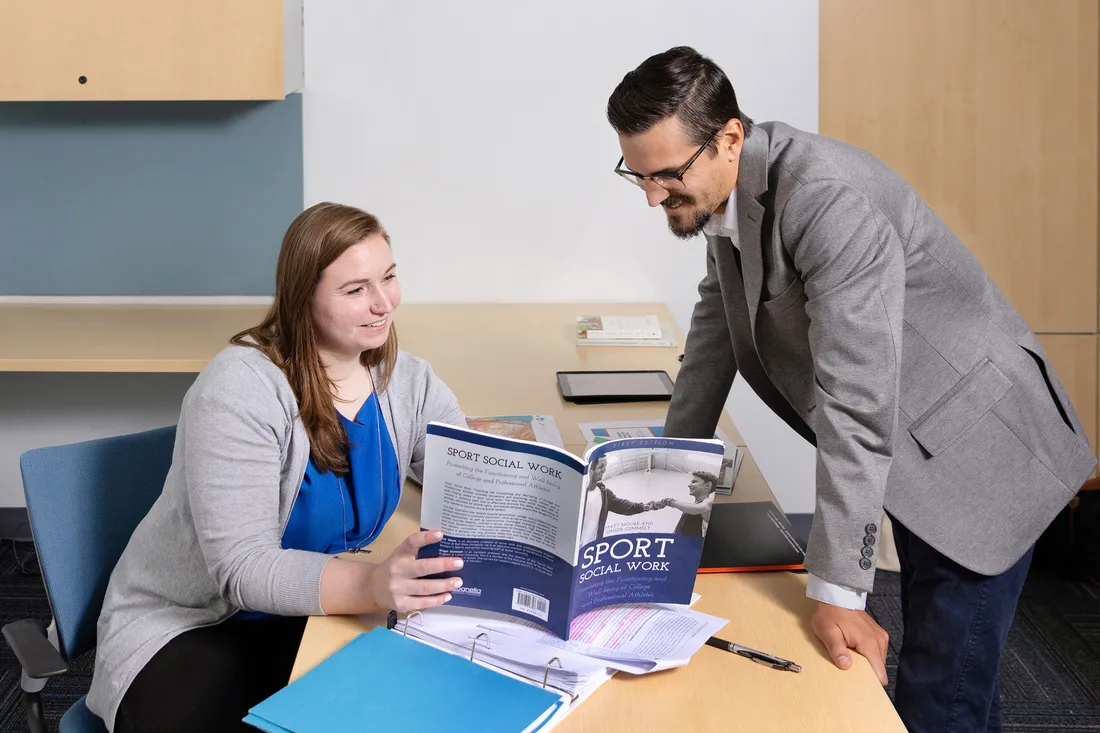 Professor and graduate student look at book together in classroom.