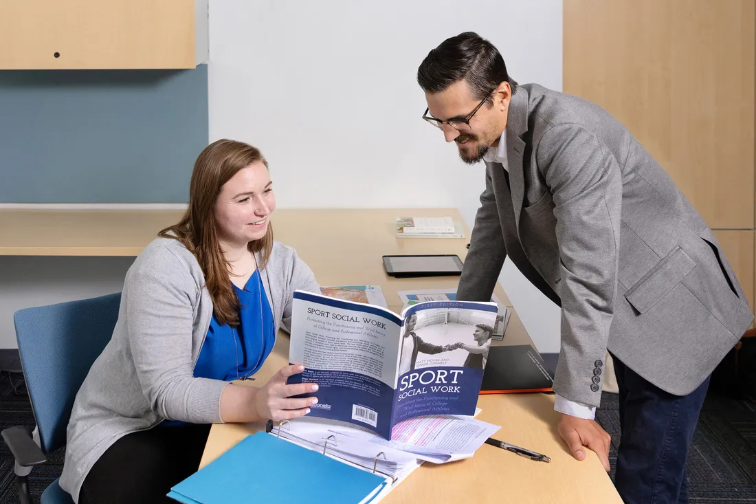 Professor and graduate student look at book together in a classroom.