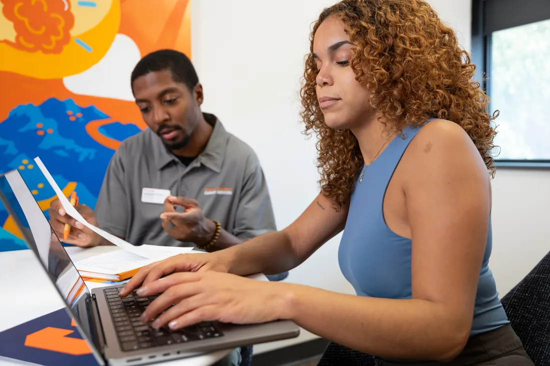 A female student typing on a computer with a male person talking to her.