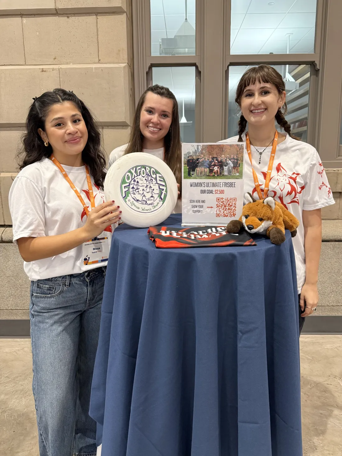 Three women standing behind a table and smiling while holding a flyer and a frisbee disc.