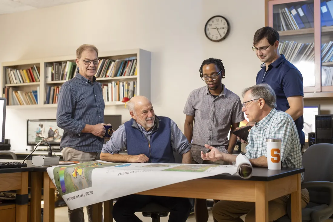 Group of faculty and students sitting and standing around a table discussing research.