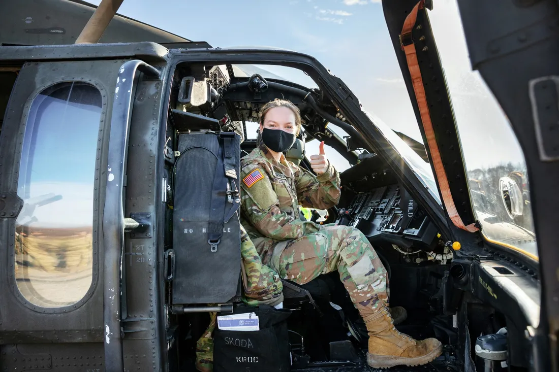 Woman in military fatigues sitting in front of vehicle giving a thumbs up.