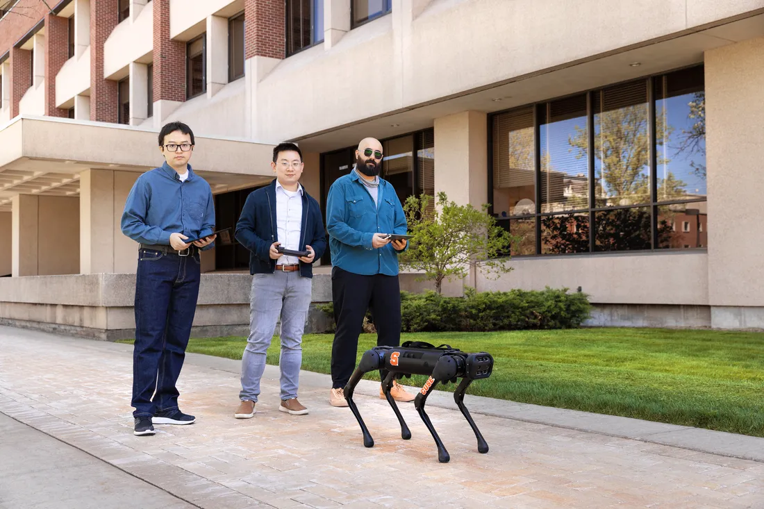 Three men outside controlling a robotic dog with remotes.