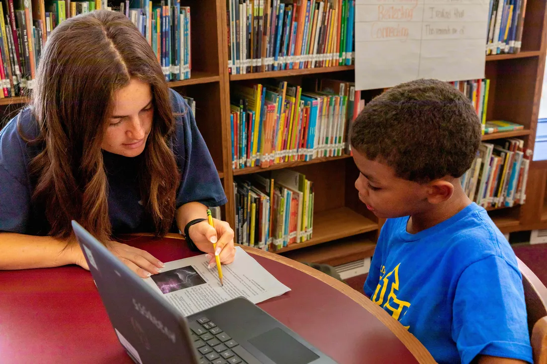 Person reading a paper to elementary age student at a table with a laptop.