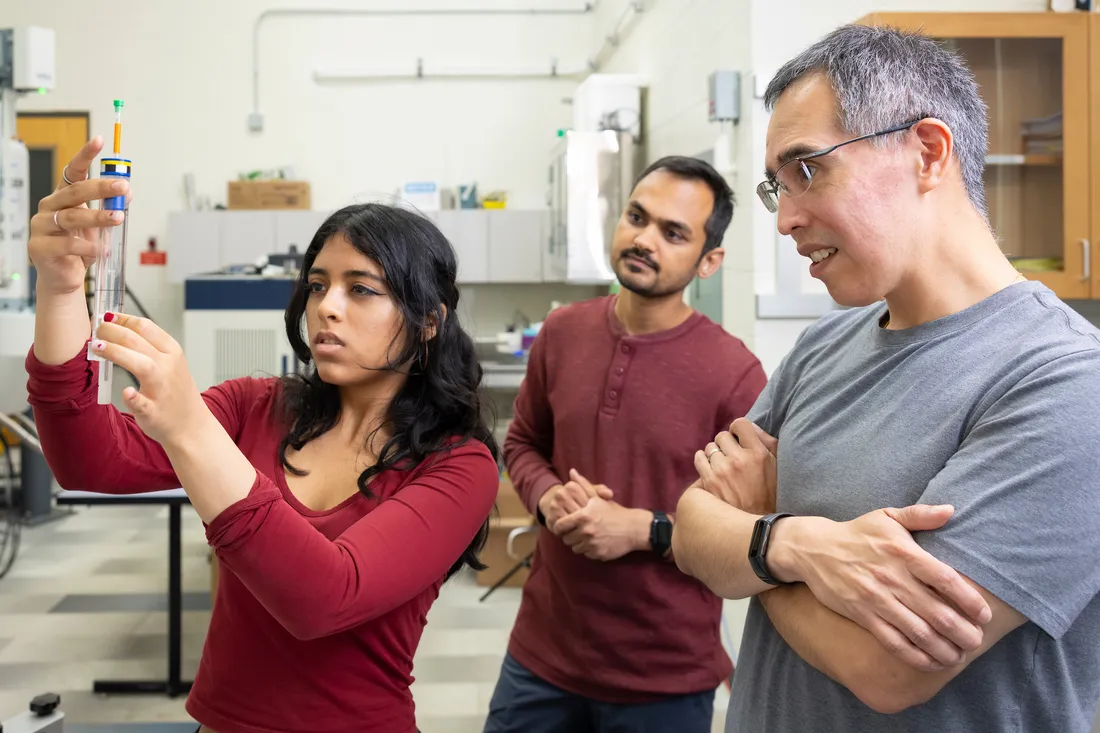 A student doing research in a lab with another student and professor looking on.