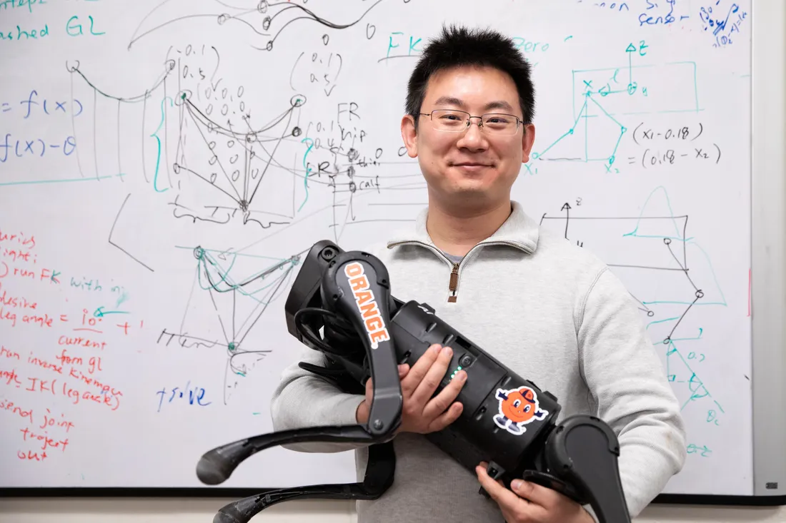 Portrait of Gan with one of his quadruped robots in front of a whiteboard covered with math.