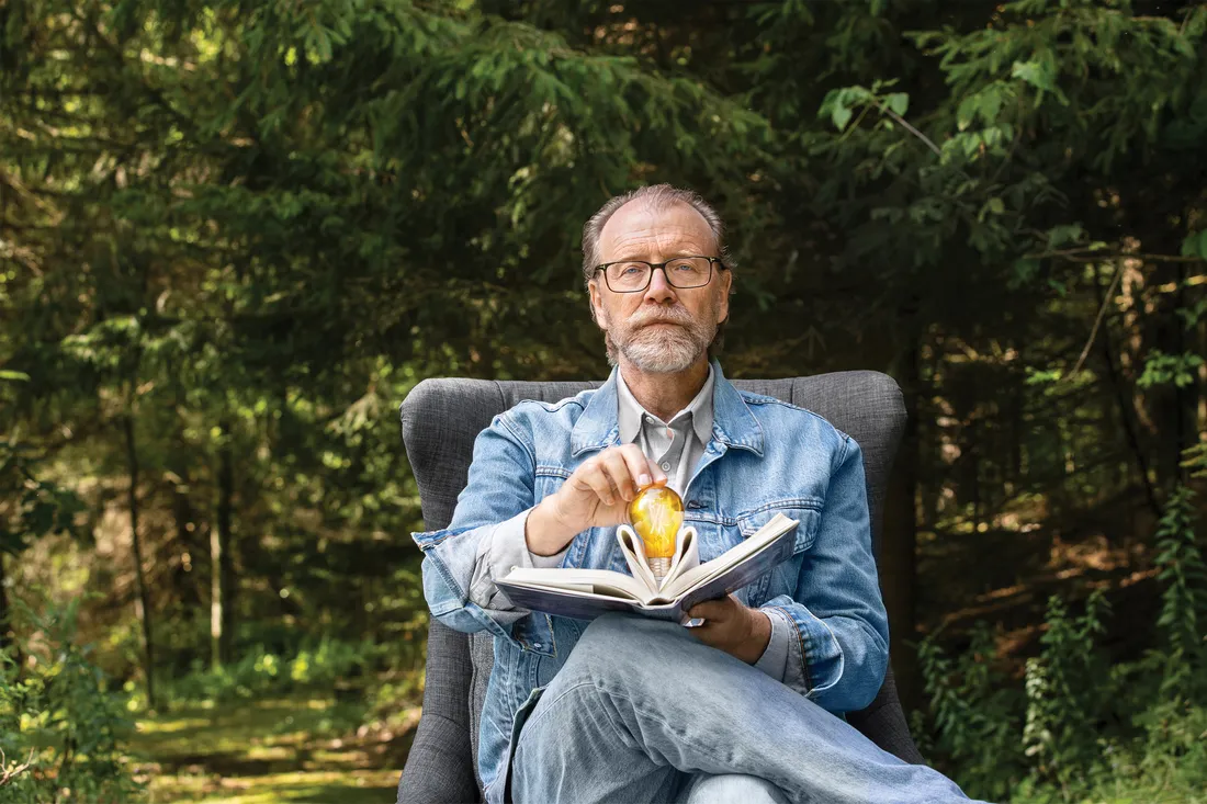 Portrait of George sitting in a chair outside, holding an open book and lightbulb in his hands.