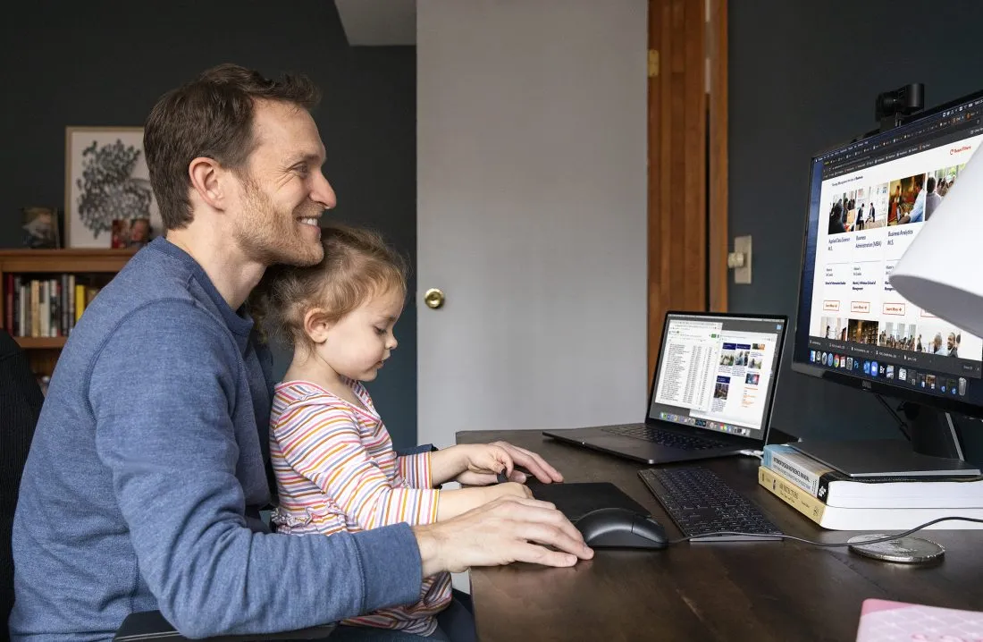 Online student Ryan Raymond sits at his desk in his home office with his daughter on his lap.