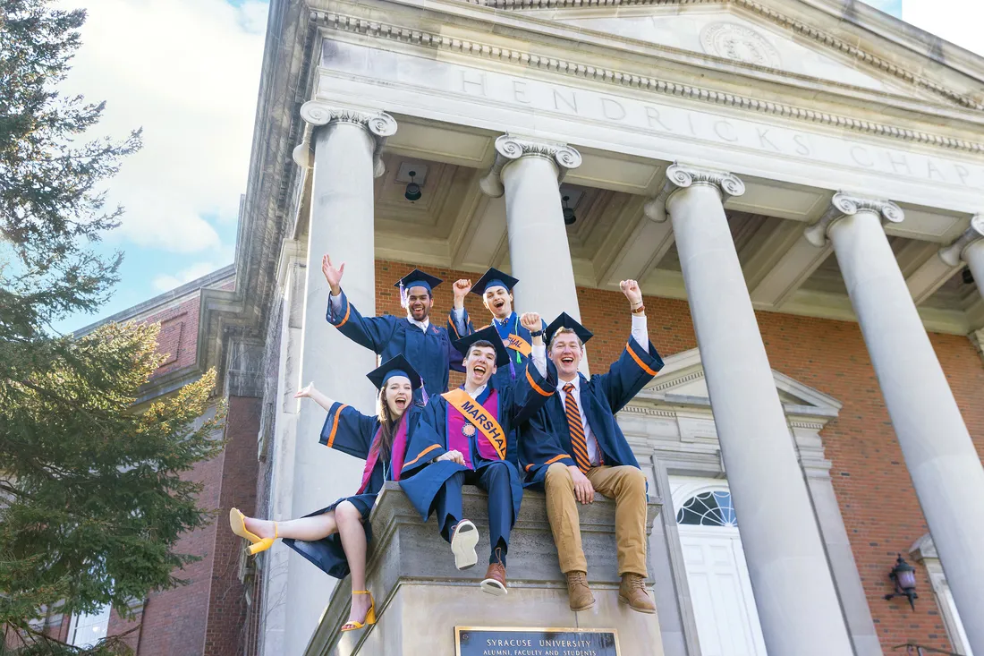 Students sit on steps of building in commencement regalia celebrating graduation.