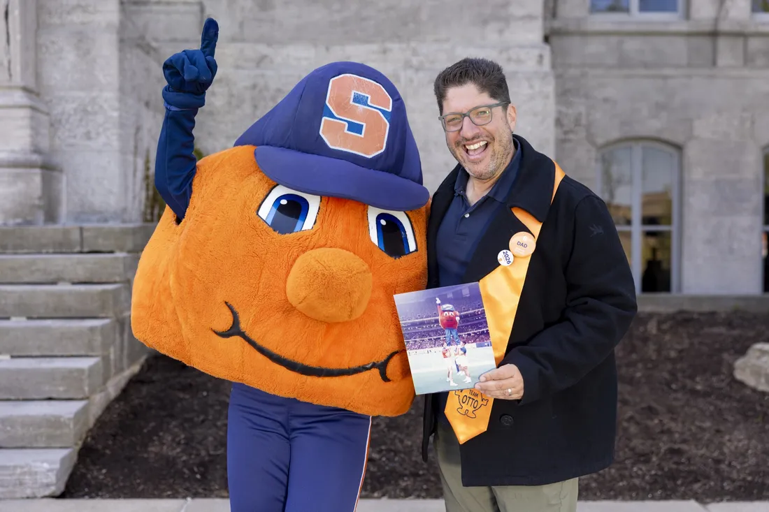 Alumnus Mitch Messinger posing with Otto the Orange and holding an old photograph of himself dressed in the Otto suit.