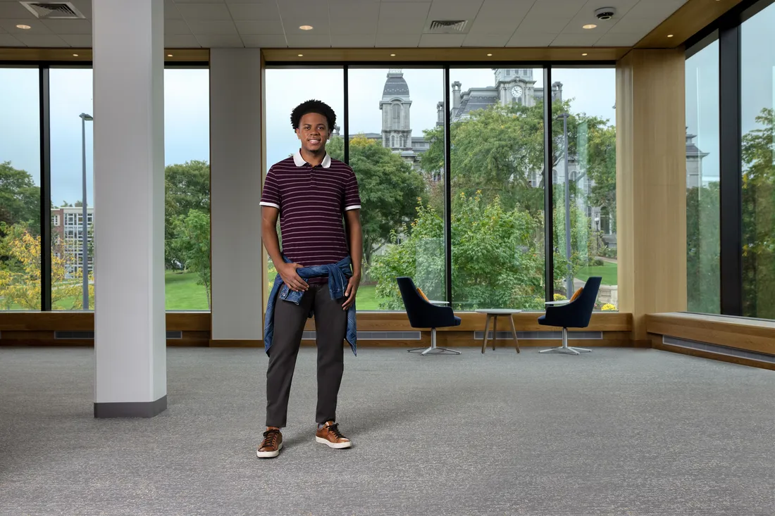 Wide portrait of David Barbier Jr. standing in front of windows, smiling.