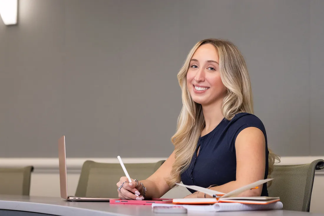Smiling student with long blonde hair writes with a stylus at a desk with a laptop and notebook.