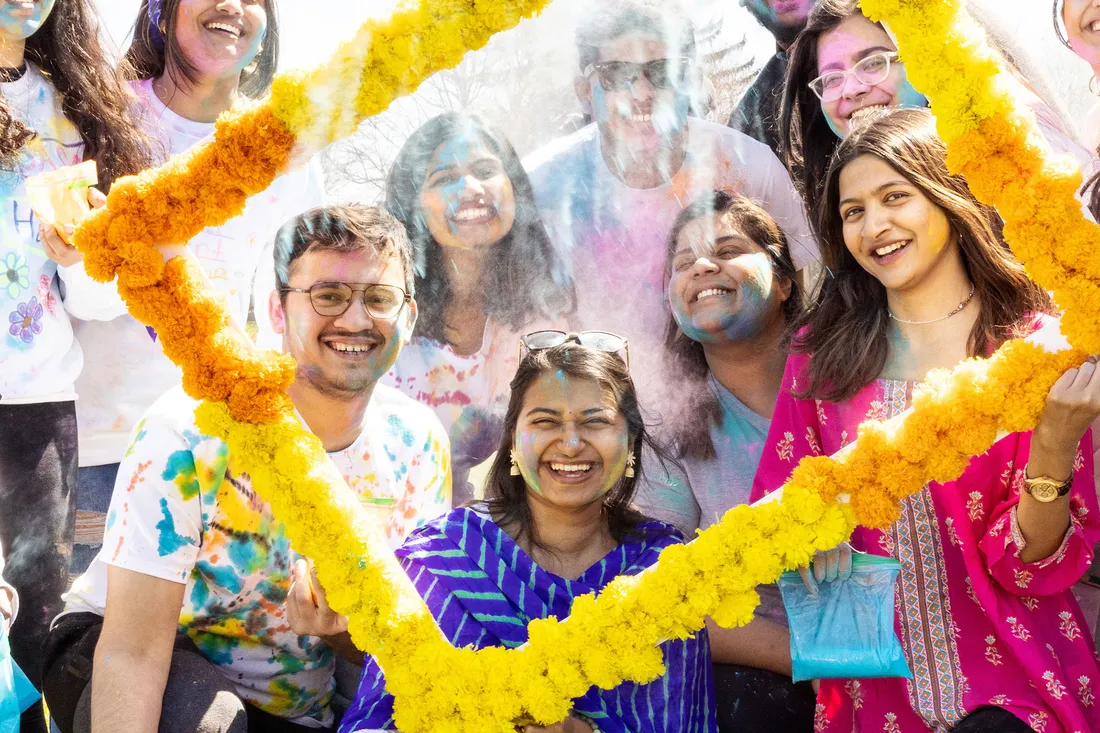 Students wearing colorful clothing and throwing colorful powder pose for a photo inside of a yellow and gold flower frame.