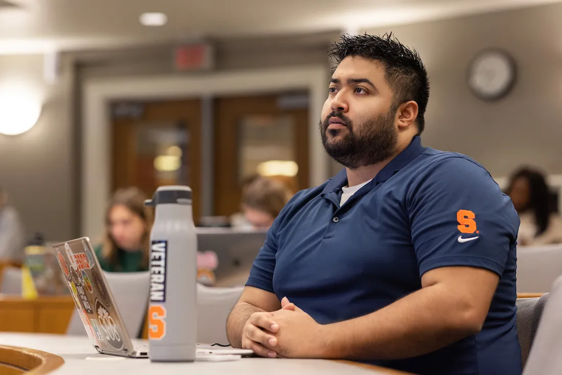 Man sitting at desk in classroom with laptop and water bottle.