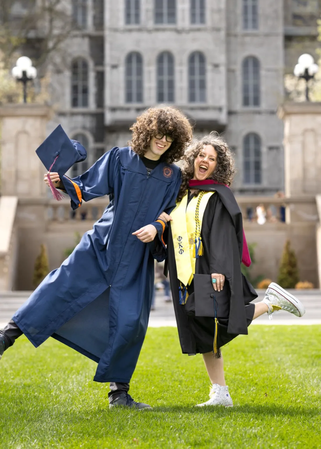 Dara Harper '26 and Rio Harper '26 smiling and jumping in their caps and gowns outside of the Hall of Languages.