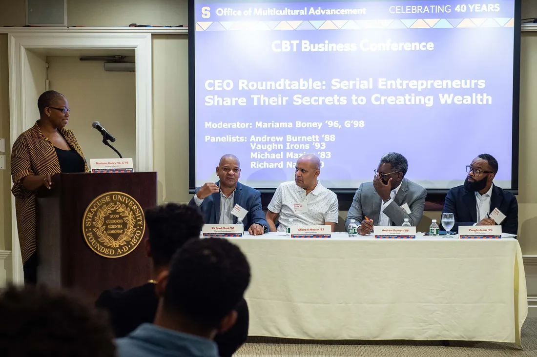 Mariama Boney standing at a podium, moderating an alumni panel discussion of four participants.