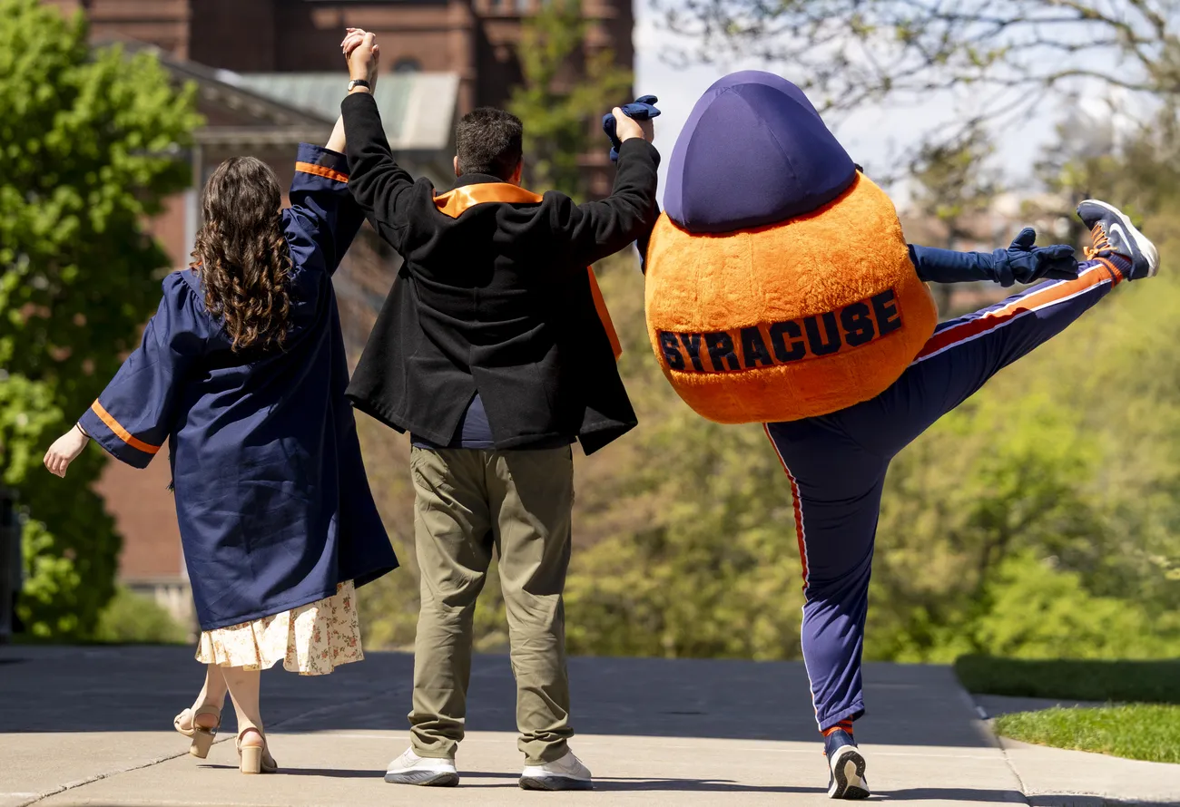 Sarah Jacoby '26, Mitch Messinger ’92, G’93 and Otto the Orange facing away from the camera and raising their arms up in celebration.