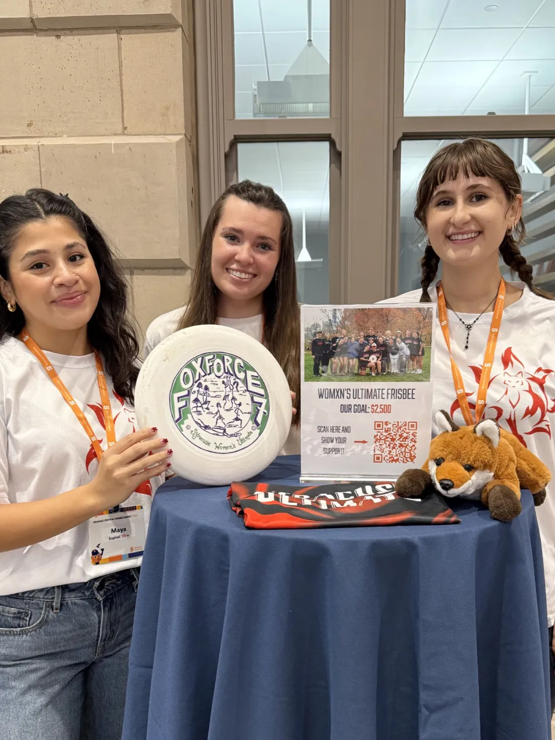 Three members of the Fox Force Seven ultimate frisbee team standing at a table holding a frisbee and flyer.