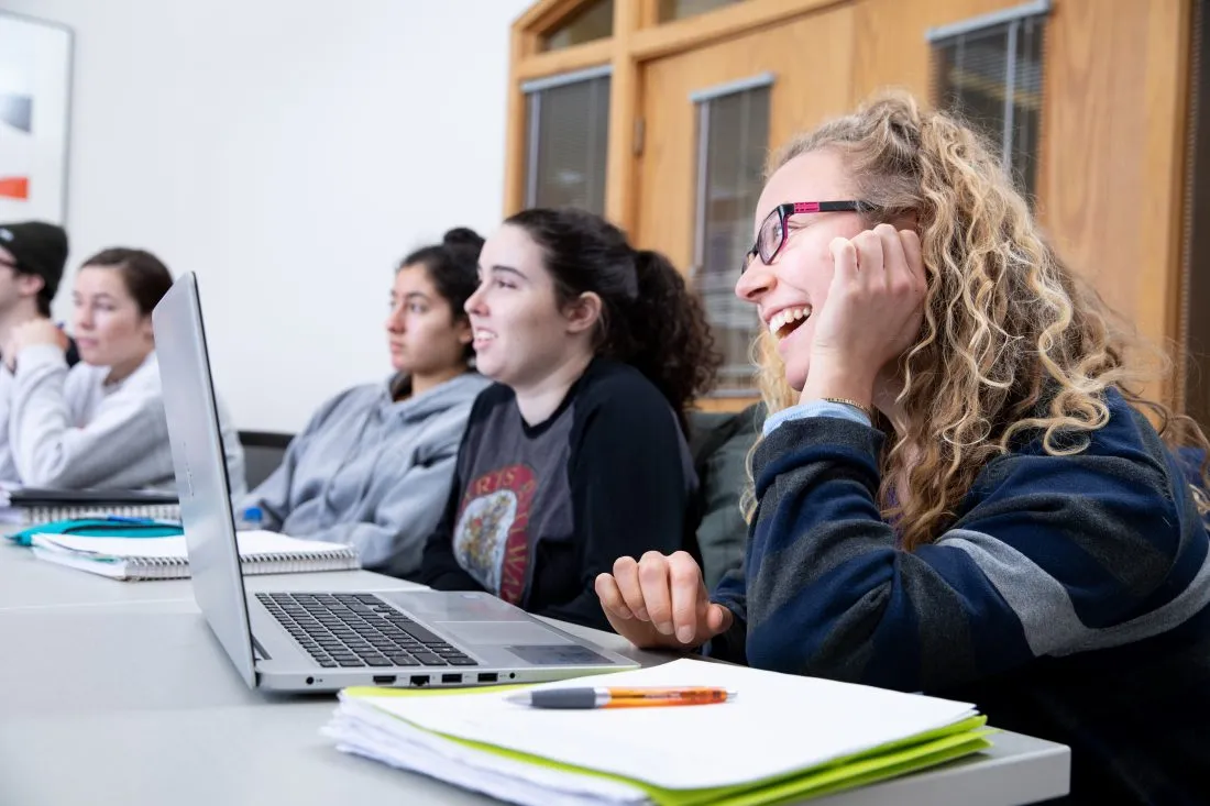 Students smiling with laptops open in front of them while sitting in a classroom.