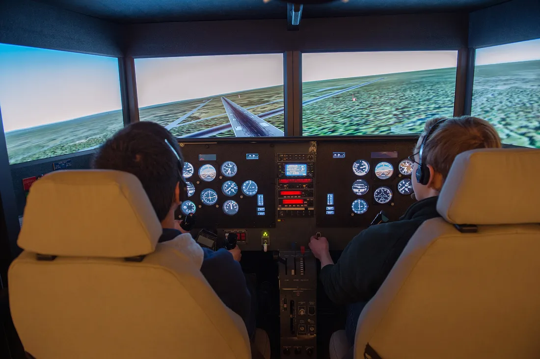 View from behind two students as they engage in the cockpit of an advanced flight simulator.