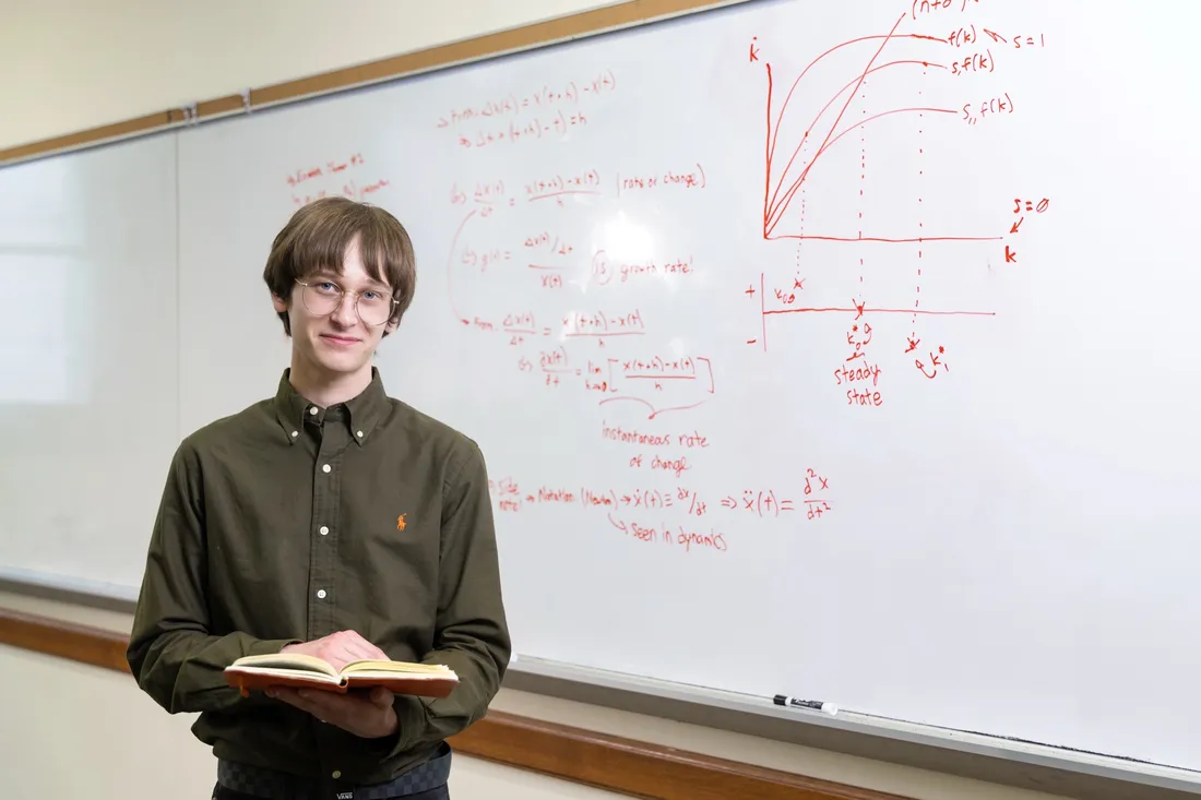 Rider Sulikowski '26 standing in a classroom in front of a whiteboard with graphs and formulas written on it.