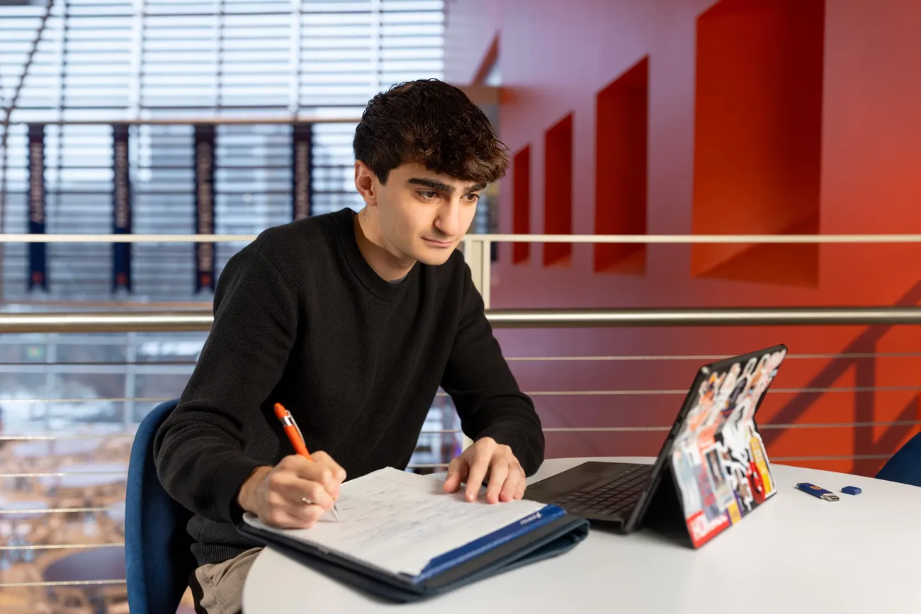 Person sitting at table looking at laptop and writing in notebook.