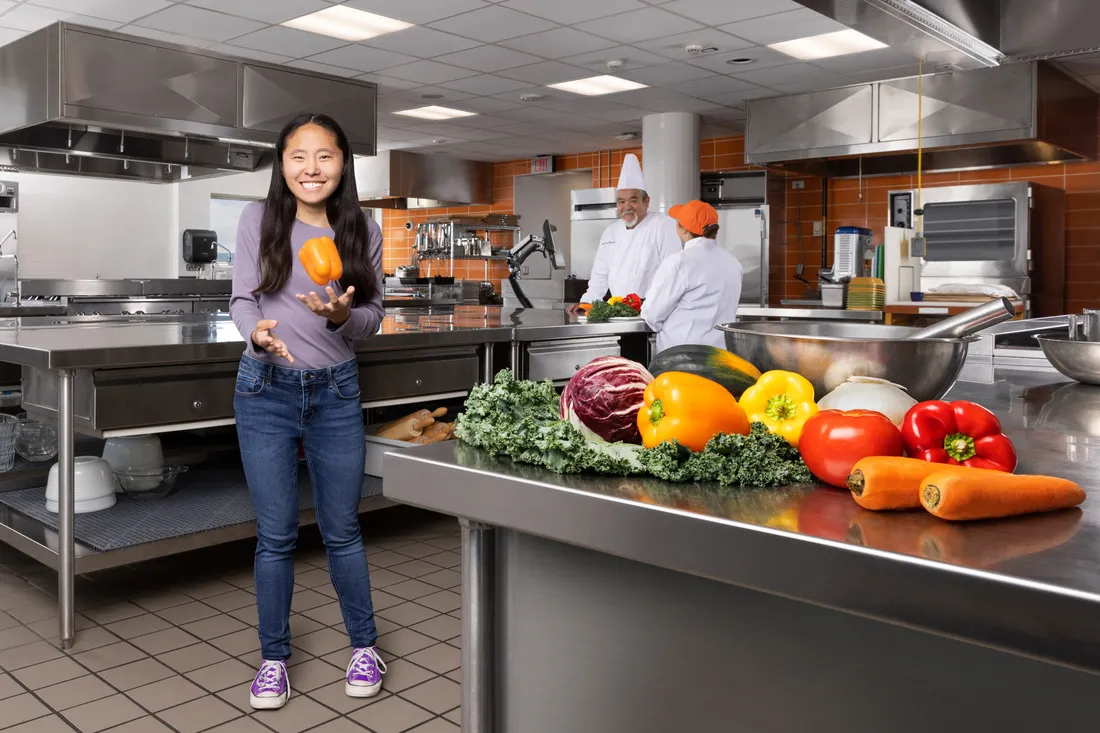Mackenzie Gleason standing in a test kitchen, catching a bell pepper midair.