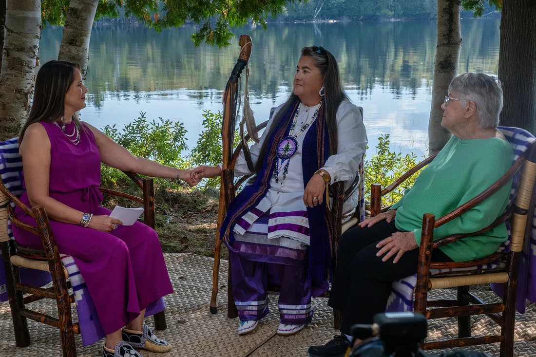 Three women sitting in wooden chairs by a body of water.