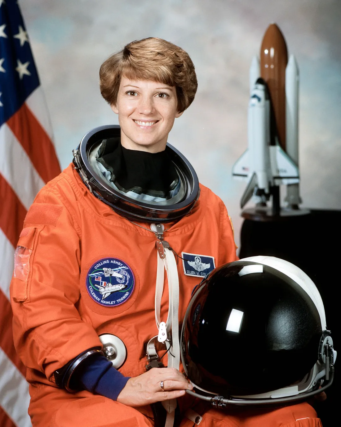 Portrait of Eileen Collins in an orange astronaut suit with her helmet on her knee.