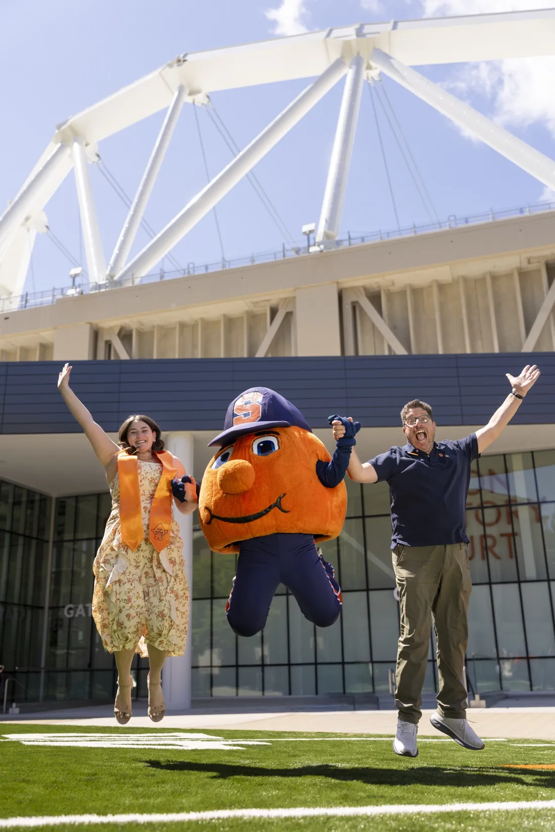 Sarah Jacoby '26, Otto the Orange, and Mitch Messinger ’92, G’93 jumping hand in hand outside of the JMA Wireless Dome.