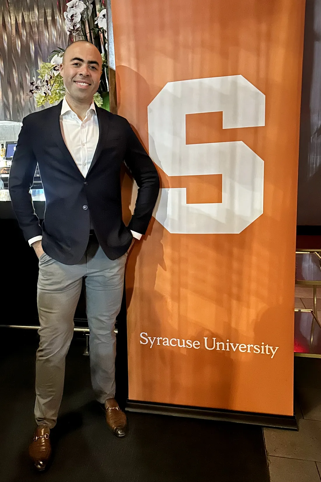 Person standing and smiling in front of the logo for Syracuse University.