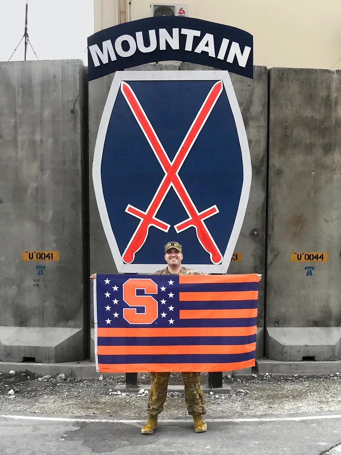 Ryan Marquette holds a Syracuse University flag in front of Army National Guard seal.